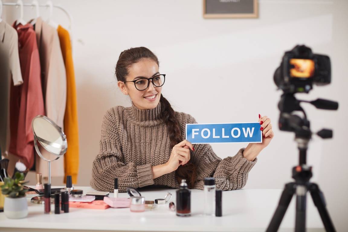 A portrait of a young woman holding a sign with "FOLLOW" on it while filming video.