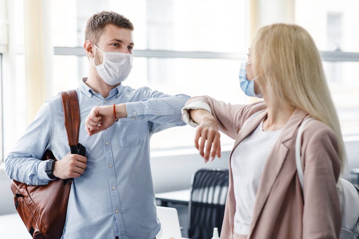 A man and woman, both in protective masks, touch elbows as a greeting as they enter an office.