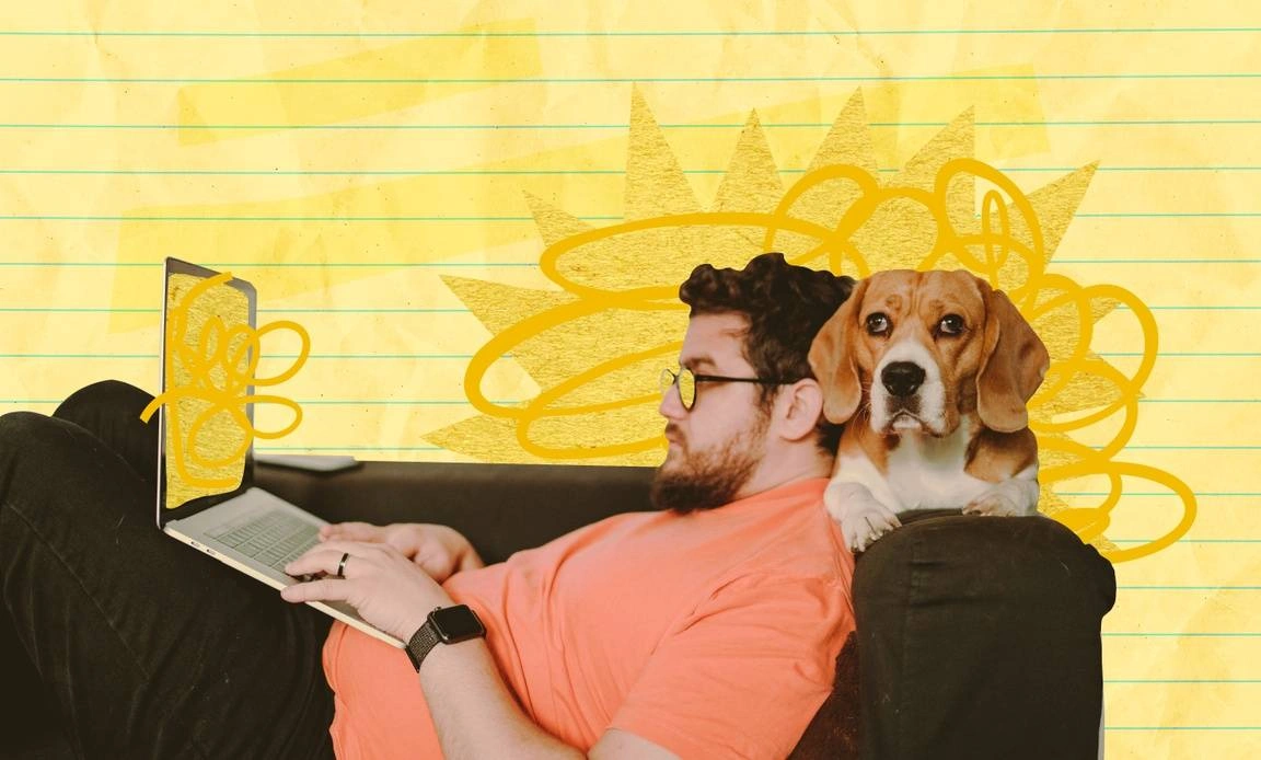 Man working on a laptop while laying on the couch next to his dog.