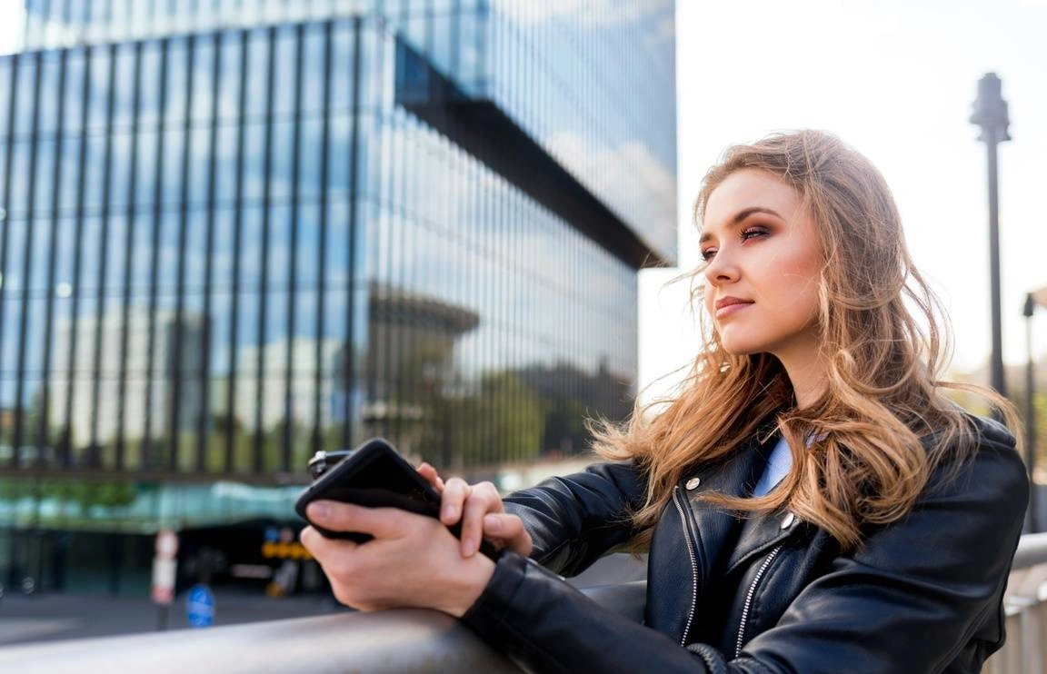 A woman holding her phone looks off into the distance of a modern-looking city.