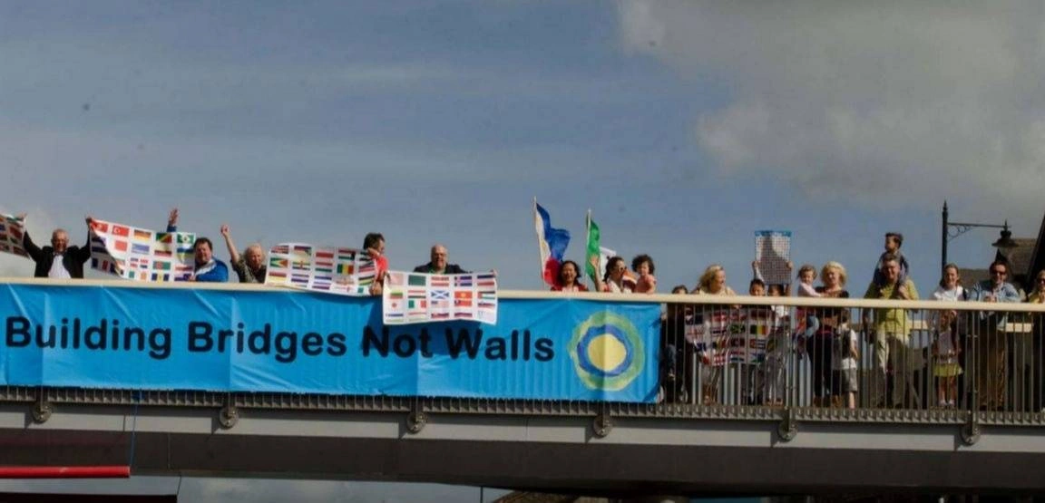 A crowd of people are standing on a bridge with a blue banner in front of them that says, "building bridges not walls."