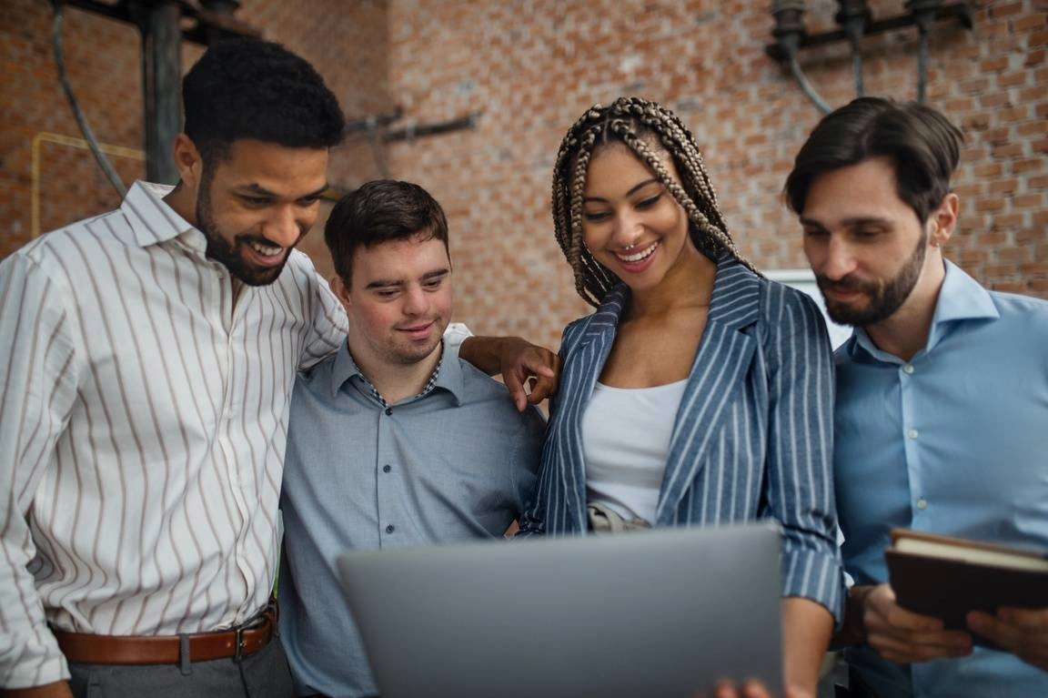Four different young employees stand together in front of a laptop.