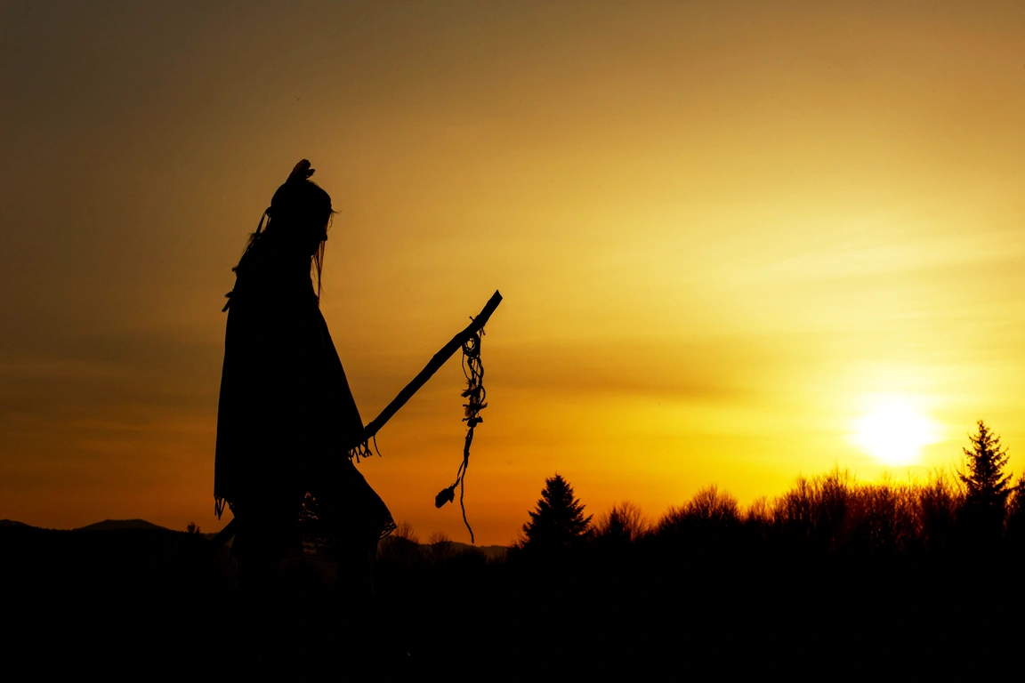 The silhouette of an Indigenous shaman holding a pikestaff with a background of the sun setting over the mountains.