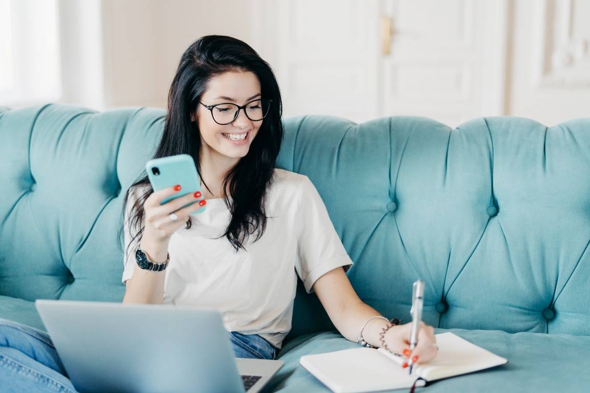 A young woman on her laptop and mobile phone, writing into a notebook