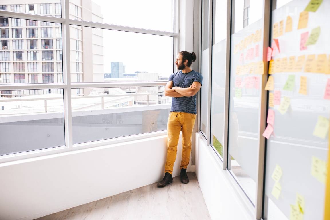 A young man with a beard and a man bun stands in front of a large glass window overlooking a city. His arms are crossed in contemplation. Behind him, a whiteboard type wall is lined with sticky notes.
