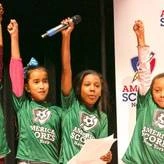 four young female students with fists raised in the air onstage at a NYC youth poetry slam