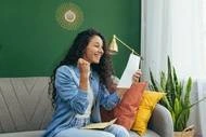 A photograph of a woman with dark curly hair excitedly looking at a piece of paper while sitting on a grey couch.