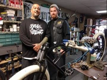 Two men pose with a chopper bicycle they are fixing.