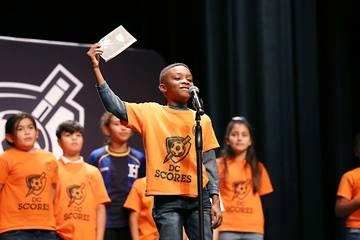 young Black boy speaking into a mic with his teammates on stage behind him