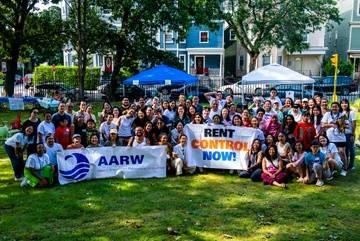 photo of a group of people, mostly Asian American, standing and smiling with banners that read "AARW" and "Rent Control Now"
