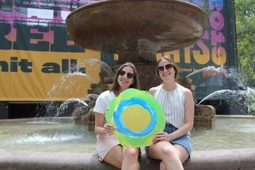 Two women sitting at a fountain hold the Idealist logo between them.