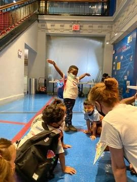 5 year old dark skinned boy wearing a Dance Project t-shirt acts out the movements of the book being read by Executive Director. Other small children are sitting nearby listening to book