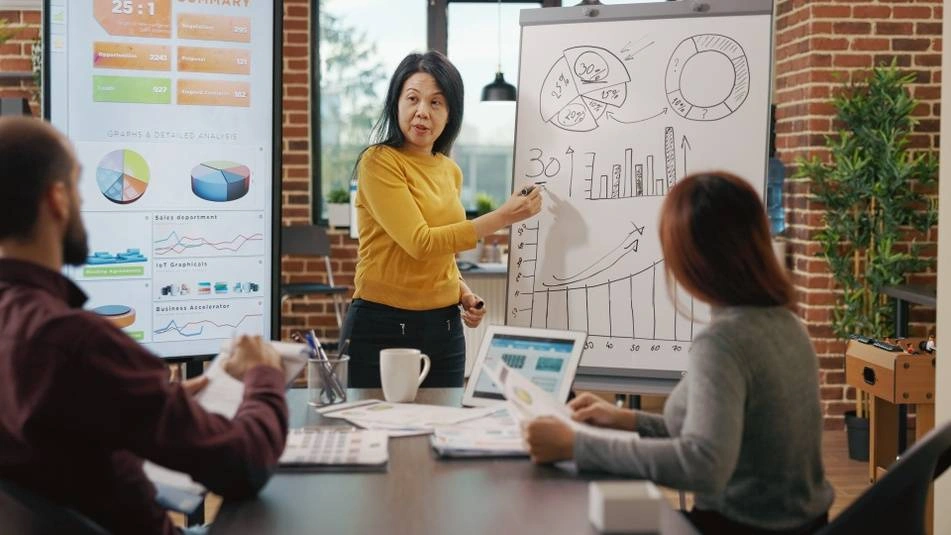 An Asian woman draws on a whiteboard during a meeting with another woman and a white man.