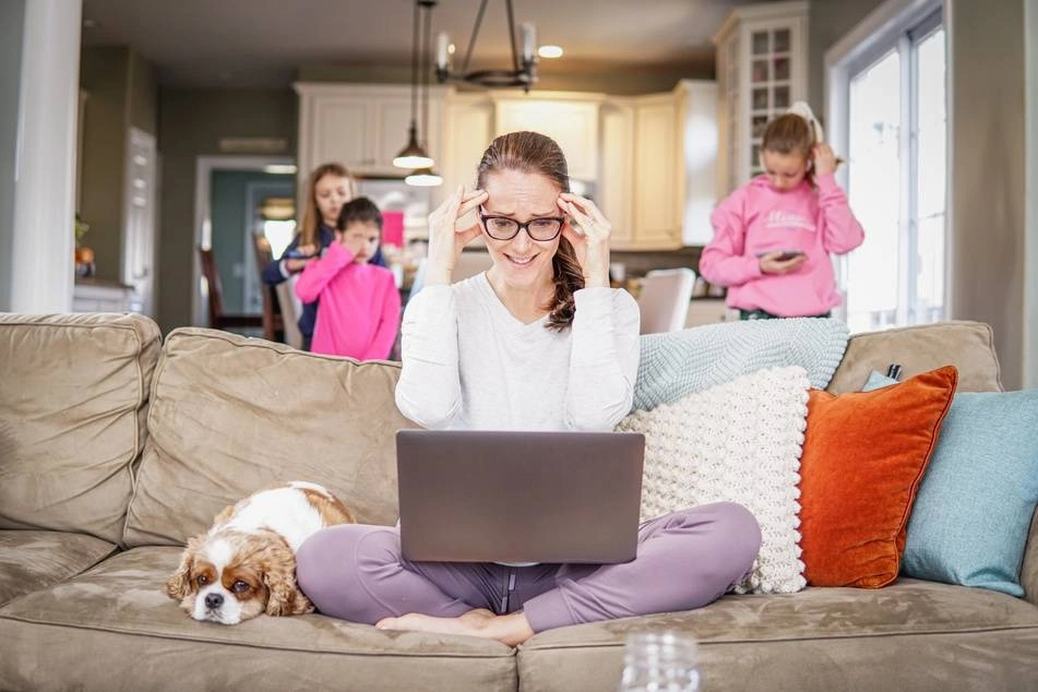 An exasperated mom leans over a laptop with her children behind her.
