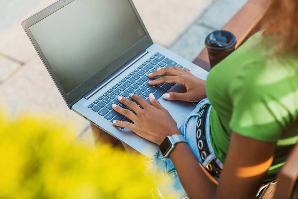 A photograph of a Black woman sitting on a bench outside with her laptop on her lap as she drinks a cup of coffee.
