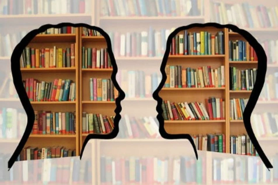 El perfil de dos personas una frente a otra y en el fondo libros en una biblioteca