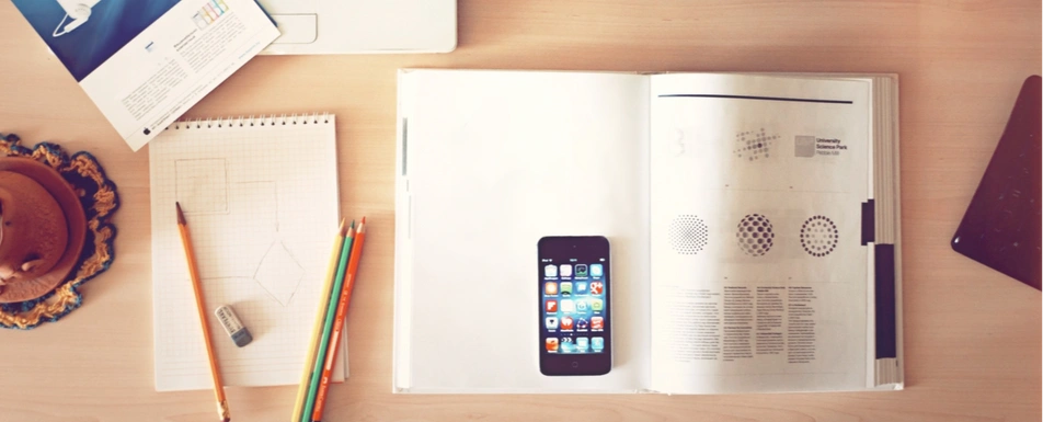 A work desk with an open book, a smartphone and a notepad with writing utensils.