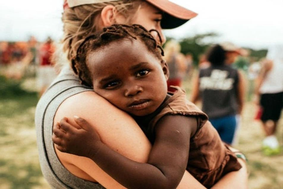 Voluntaria blanca y rubia cargando en brazos a bebé negro de escasos recursos