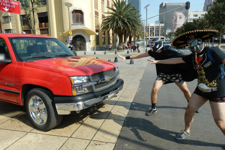 Dos hombres disfrazados con máscaras del Santo, capas y sombrero mexicano frenando un carro