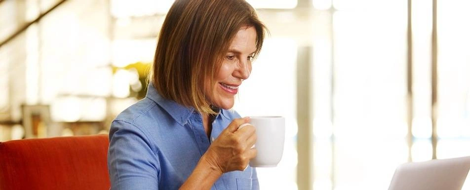 A woman sits at a laptop drinking coffee.