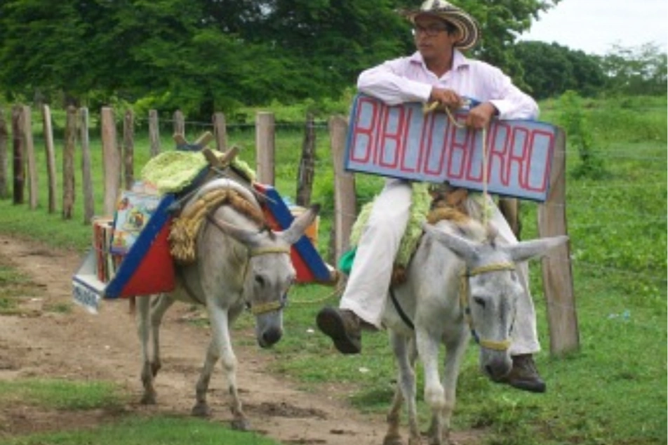 Persona montando un burro y llevando libros hacia los pueblos, con un letrero que dice Biblioburro