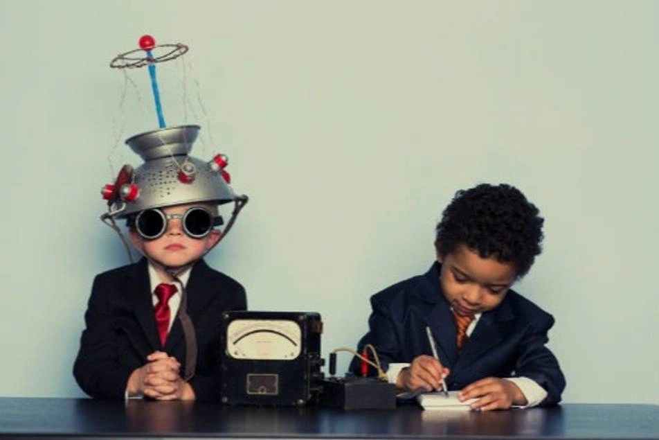 Dos niños disfrazados con traje y corbata. Uno con una olla en la cabeza simulando un científico y el otro escribiendo como un oficinista.