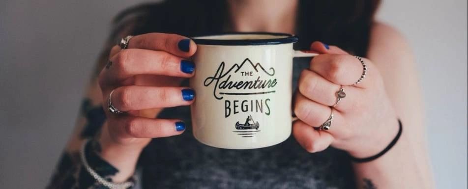 A woman with blue fingernails holds a mug that says, "The adventure begins."